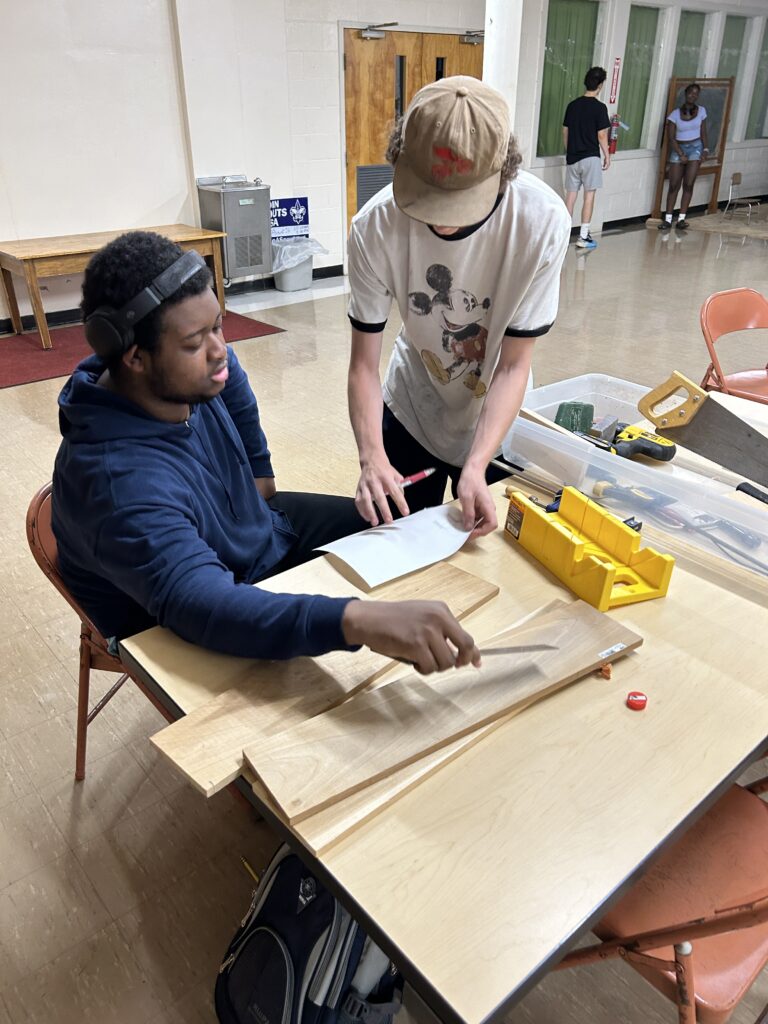 One high school student standing next to a seated high school student, looking at a drawing, with wood and tools nearby.
